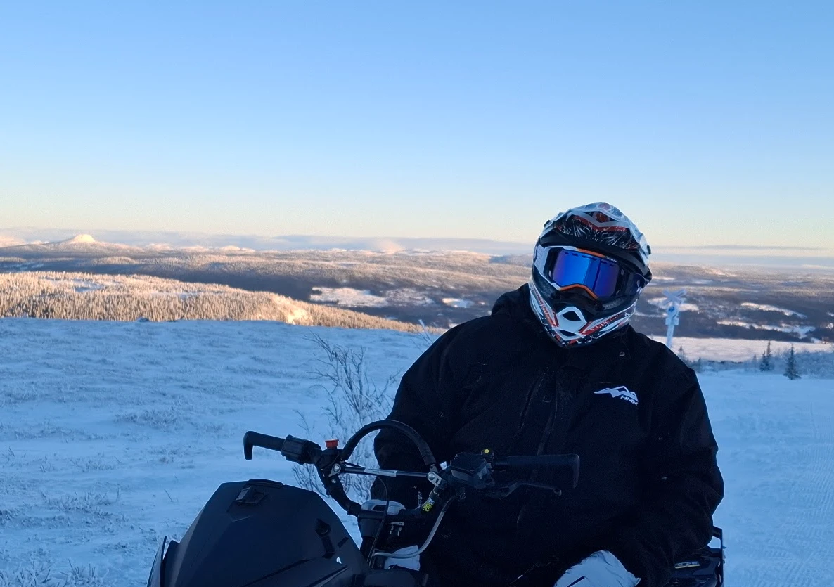 Skoterförare med hjälm på fjälltopp, milsvid utsikt över snöklädda fjäll